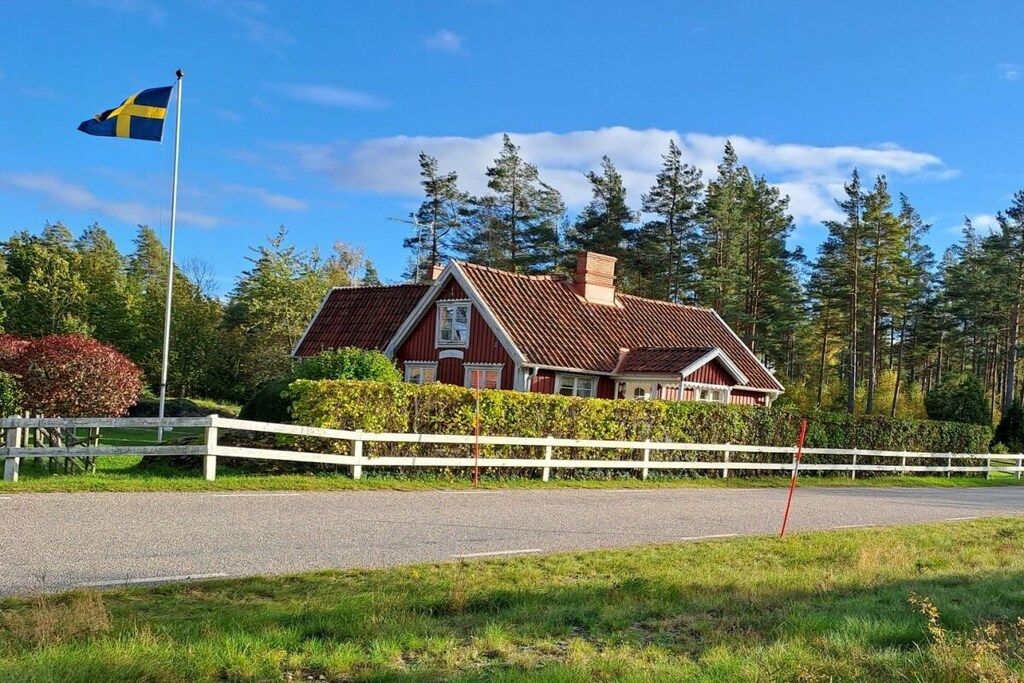 A traditional red-roofed house with a white fence, a flagpole with a flag, and a forested background.