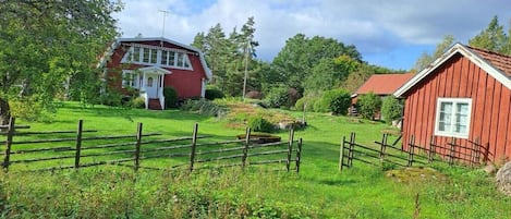 A red wooden house with a white door and a small porch, surrounded by a wooden fence and lush greenery.