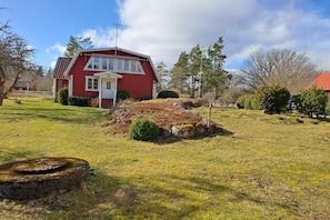 A red house with a white door and windows, surrounded by a grassy yard with a stone structure and trees. - Holiday home with 1 bedroom (Söderakra)