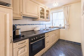 A kitchen with light wooden cabinets, a black stove, and a window with curtains. - Holiday apartment in Schönwald with barbecue (Schönwald)