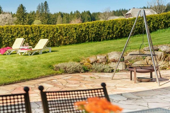 Een terras met stenen vloer, twee loungestoelen, een schommel en een vuurkorf.