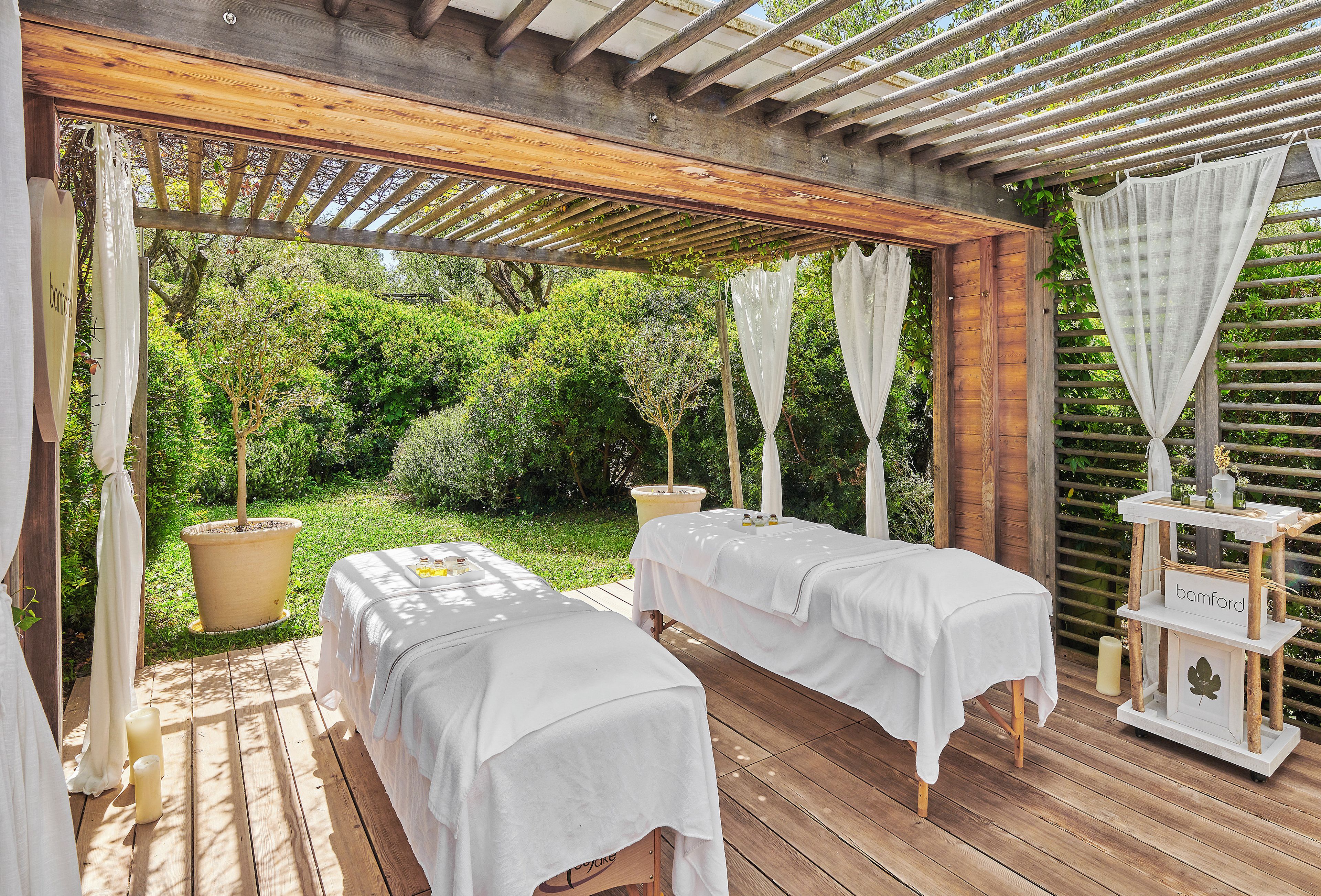 Une terrasse en bois avec deux tables de massage recouvertes de draps blancs, une pergola en bois au-dessus, et une table blanche portant une pancarte indiquant « bambord ».