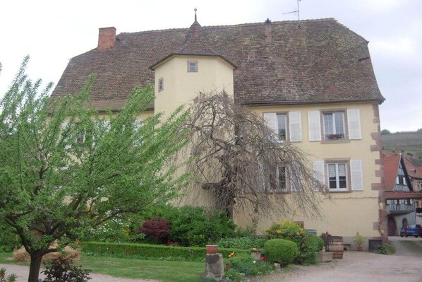 A two-story house with a sloped roof, a small balcony, and a garden with various plants and flowers. - Holiday apartment with 1 bedroom (ANDLAU)