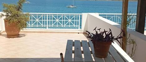 A terrace with a wooden table and benches, overlooking a body of water with boats and hills in the distance.