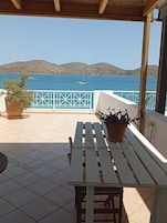 Una terraza con mesa y bancos de madera, con vistas a una masa de agua con barcos y colinas a lo lejos.