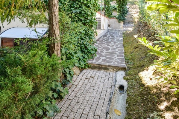 En brostenssti fører hen til en bygning med et overdækket indgangsparti, omgivet af grøn vegetation og et træ.