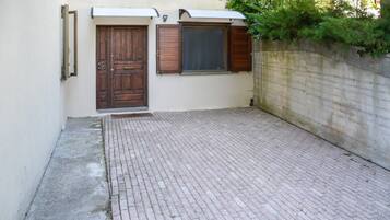 Ein gepflasterter Innenhof mit einer Holztür und Fensterläden, einer Betonmauer und einer kleinen Stufe zum Eingang.