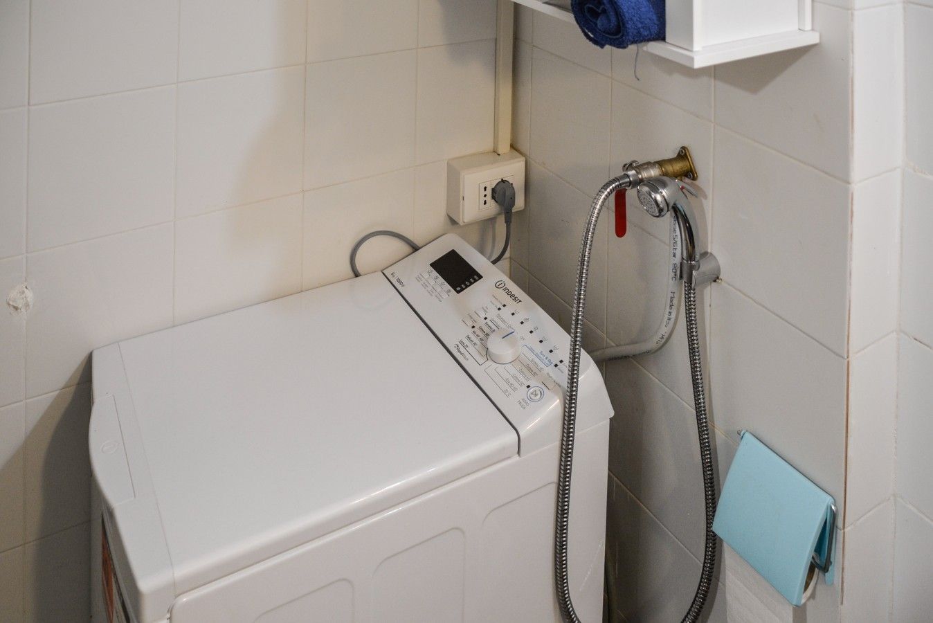 Eine weiße Waschmaschine mit Bedienfeld, einer Steckdose und einem Schlauch, der an einem Duschkopf angeschlossen ist.
