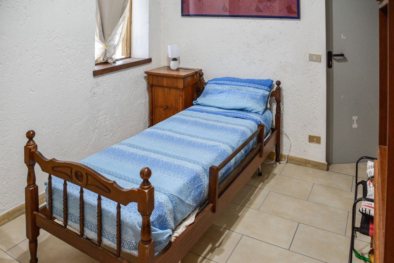 Cama de solteiro com estrutura de madeira e colcha listrada azul em quarto com paredes brancas e chão de azulejos.
