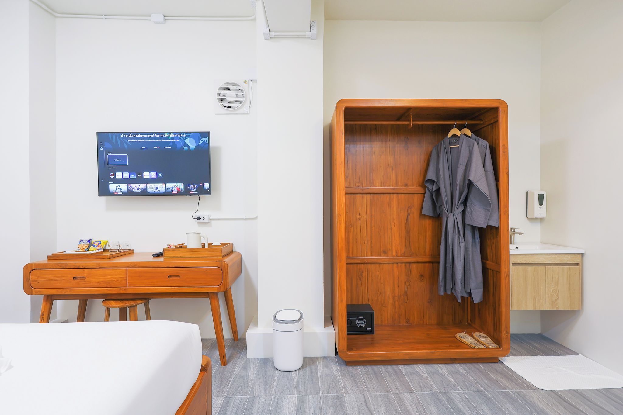 A hotel room with a wooden desk, a television mounted on the wall, a wardrobe with a robe hanging, and a small bathroom area.