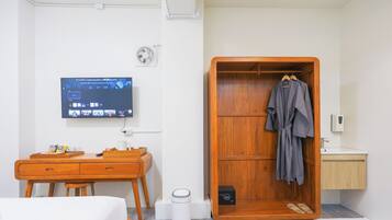 A hotel room with a wooden desk, a television mounted on the wall, a wardrobe with a robe hanging, and a small bathroom area.