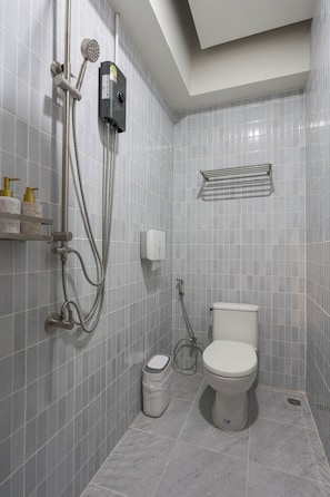 Casa de banho com sanita, chuveiro com duche manual, toalheiro e um cesto de lixo.
