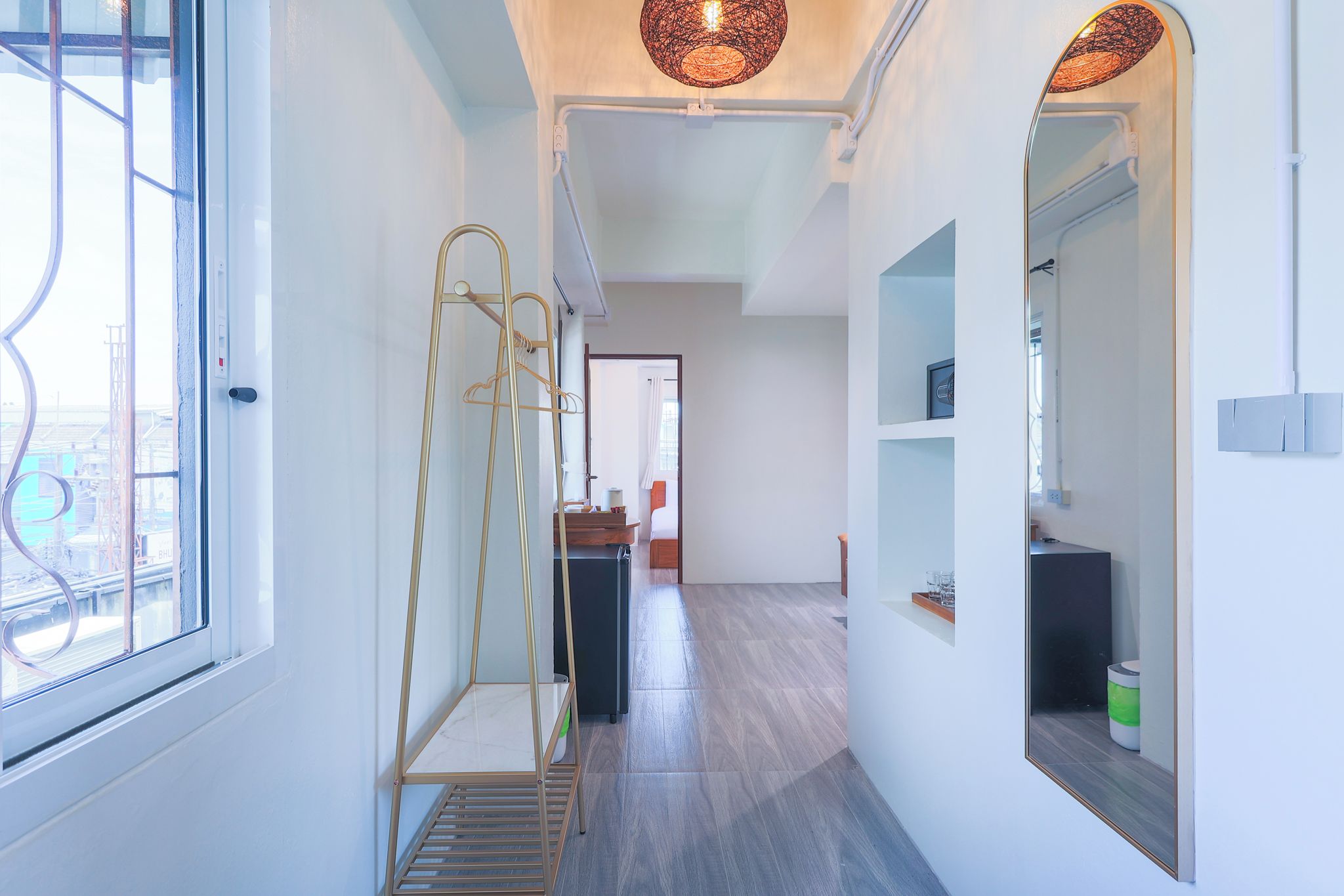 A hallway with a window, a metal rack, a mirror, and a door leading to another room.