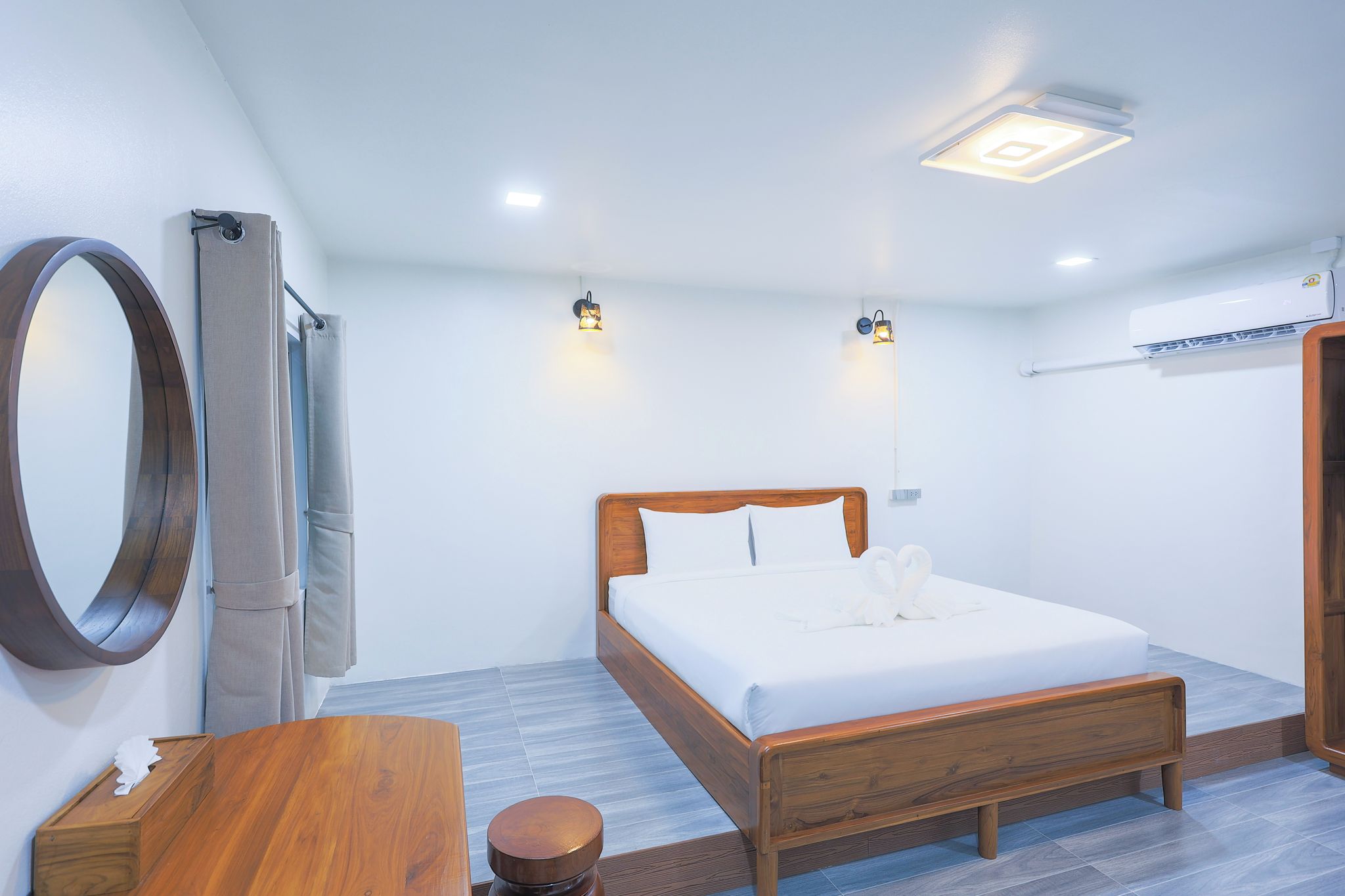 A hotel room with a wooden bed, a round mirror, a wooden side table, and a small stool.
