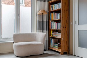 A room with a white textured armchair, a wooden bookshelf filled with books, a floor lamp, and a window with curtains. - Lovely house in Zoutelande for 8 persons - Duinweg 11 (Zoutelande)