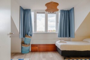 A bedroom with a bed, a window with curtains, a radiator, and a hanging light fixture. - Lovely house in Zoutelande for 8 persons - Duinweg 11 (Zoutelande)