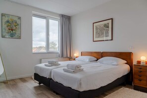 Una cama doble con dos almohadas, una mesita de noche con lámpara, un radiador y una ventana con cortinas.