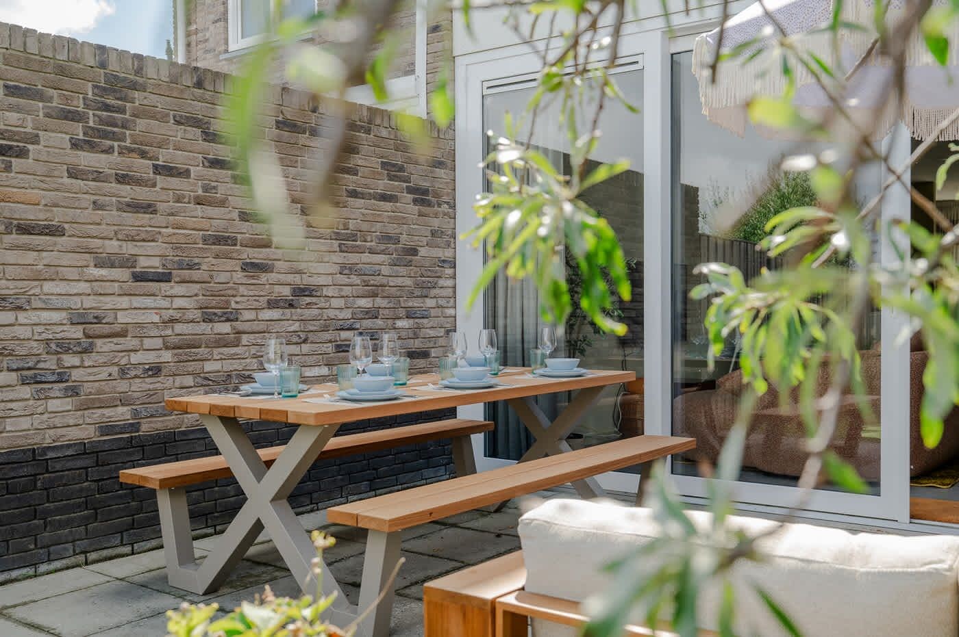 An outdoor dining area with a wooden table and benches, a brick wall, and a glass door leading to a garden.