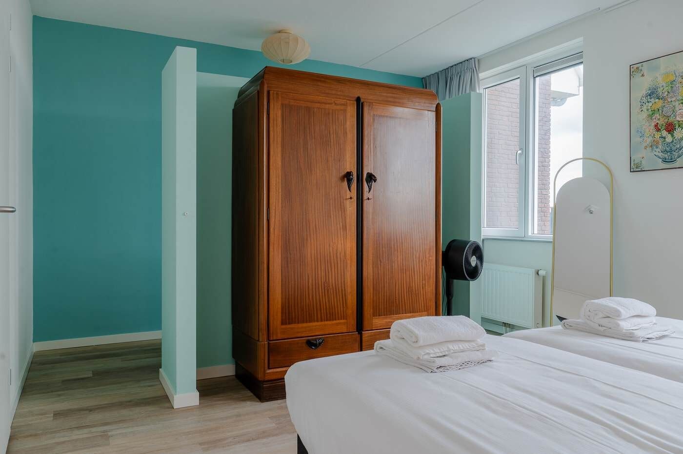 Un dormitorio con paredes verde azulado, un armario de madera, una cama con ropa blanca y una ventana con vistas.