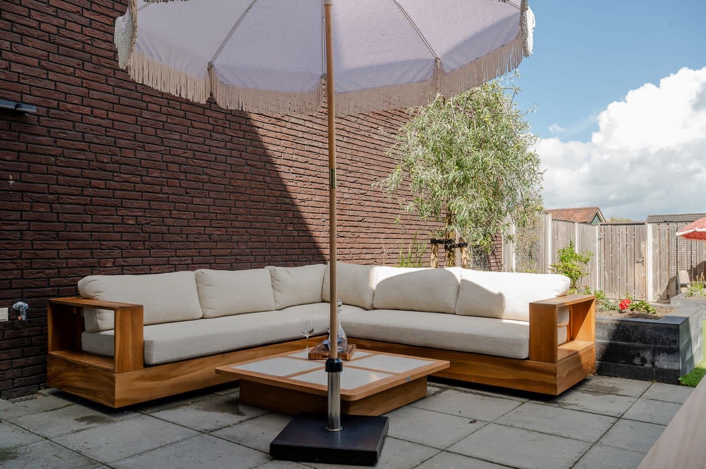 Un canapé d’angle avec des pieds en bois et une table basse assortie, placés sur une terrasse carrelée avec un mur en briques en arrière-plan.