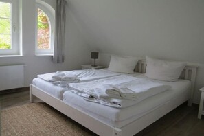 Cama doble con sábanas y toallas blancas, mesita de noche con lámpara y ventana con cortinas.