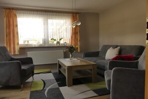 Ein Wohnzimmer mit Sofa, Sesseln, Couchtisch und einem Fenster mit Vorhängen.