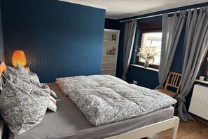 Ein Schlafzimmer mit einem Bett, einem Nachttisch, einer Lampe, einem Fenster mit Vorhängen und einer Heizung.