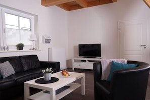A modern living room with a black leather sofa, a white coffee table with a plant, a flat-screen TV on a white stand, and a door to the right. - Cieslik holiday half-timbered house (Hollern-Twielenfleth)