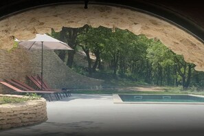 An outdoor swimming pool area with a stone wall, a bench, and a tree-lined backdrop.