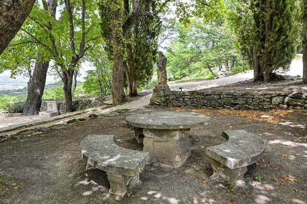 A stone bench with a statue in a park with trees and a stone wall.