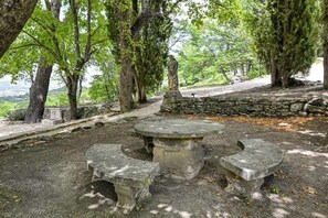 A stone bench with a table in a wooded area with trees and a stone wall.