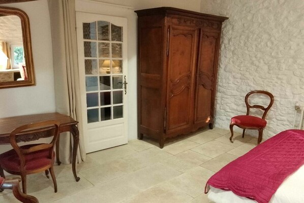A bedroom with a wooden wardrobe, a desk with a chair, a bed with a red blanket, and a white wall.