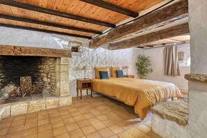 A bedroom with a stone fireplace, wooden beams, a bed with a yellow bedspread, and a tiled floor.