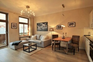 A modern living room with a sofa, a dining table with chairs, a coffee table, and a chandelier. There are framed pictures on the walls and a window with a view. - Villa Käthe "Apartment - 6b" (Kühlungsborn)