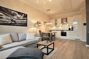 A modern living room with a sofa, coffee table, and a dining area with chairs. A kitchen with white cabinets and appliances is visible in the background. - Villa Käthe "Apartment - 6b" (Kühlungsborn)