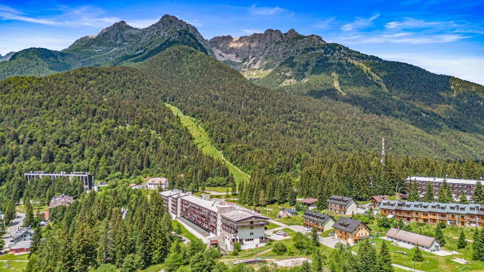 Eine gebirgige Landschaft mit einem Hotelkomplex, der aus mehreren Gebäuden besteht, darunter ein großes Hotel und kleinere Pensionen, umgeben von dichtem Wald und blauem Himmel.