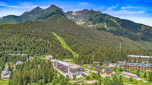 A mountainous landscape with a hotel complex featuring multiple buildings, including a large hotel and smaller guesthouses, surrounded by dense forest and clear skies. - Nevea Lux with Sauna & Kanin View (Sella Nevea)