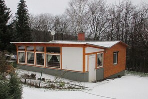 Eine kleine, eingeschossige Holzhütte mit geneigtem Dach, großen Fenstern und einem sichtbaren Schornstein.