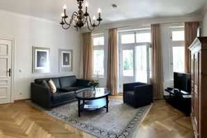 A living room with a sofa, armchairs, a coffee table, and a TV. There are large windows with curtains, a chandelier, and framed pictures on the wall. - Villa Achterkerke / upper floor south & sea (Seebad Heringsdorf)