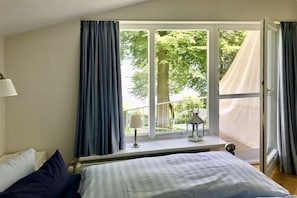A bedroom with a large window, a bed, a nightstand, and a lamp. - Villa Achterkerke / upper floor south & sea (Seebad Heringsdorf)