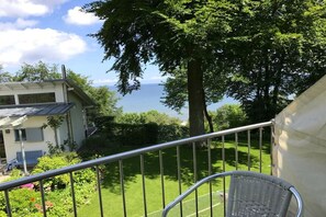 Ein Balkon mit Metallgeländer, einem Korbstuhl und Blick auf eine grüne Rasenfläche sowie ein Gebäude mit Satteldach.