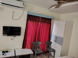 A room with a flat-screen TV mounted on the wall, a desk with a chair, a wardrobe, and a window with red curtains.
