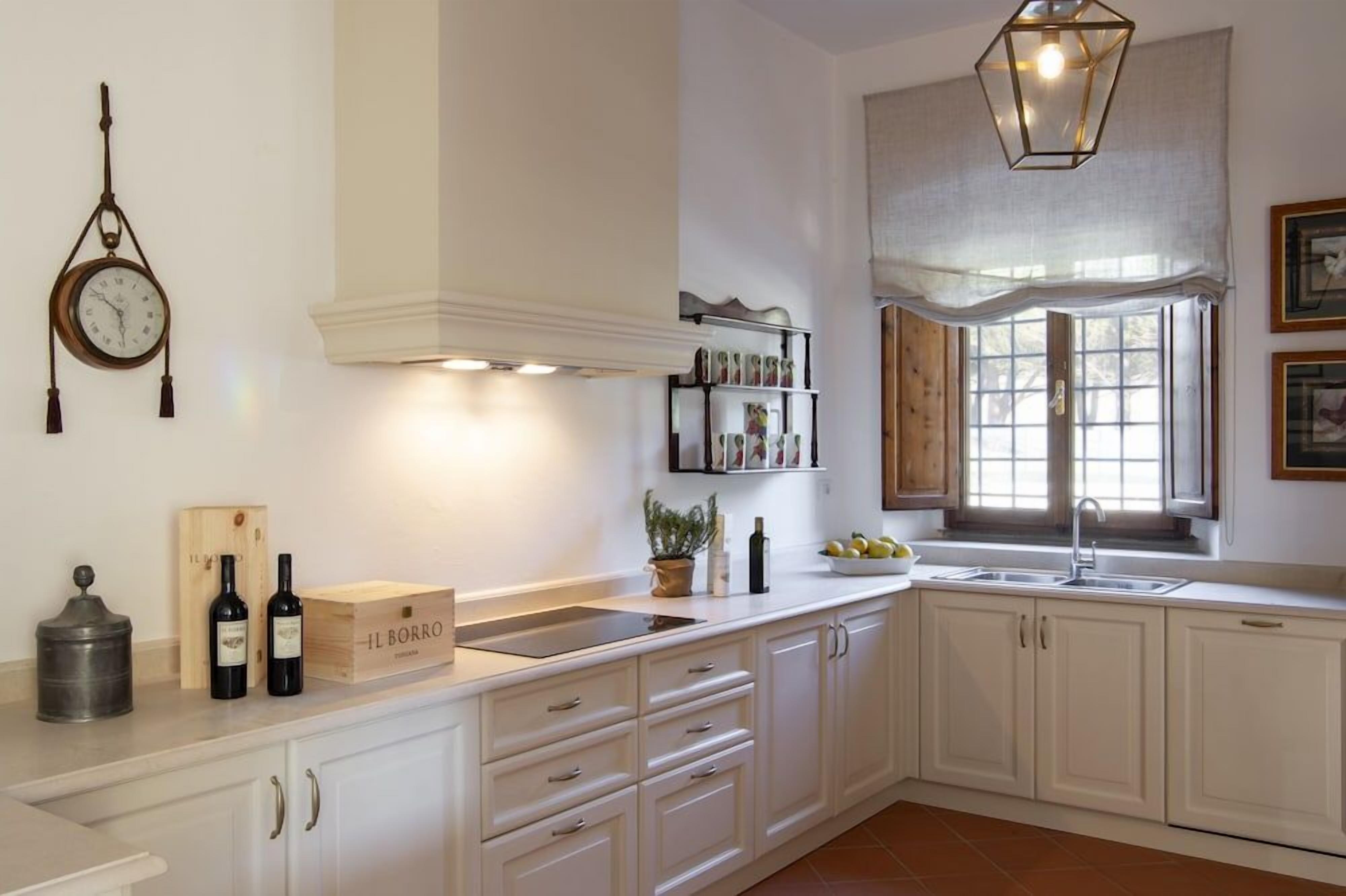 Une cuisine avec des meubles blancs, une horloge suspendue, une fenêtre donnant sur un beau paysage et une étagère remplie de bouteilles de vin.