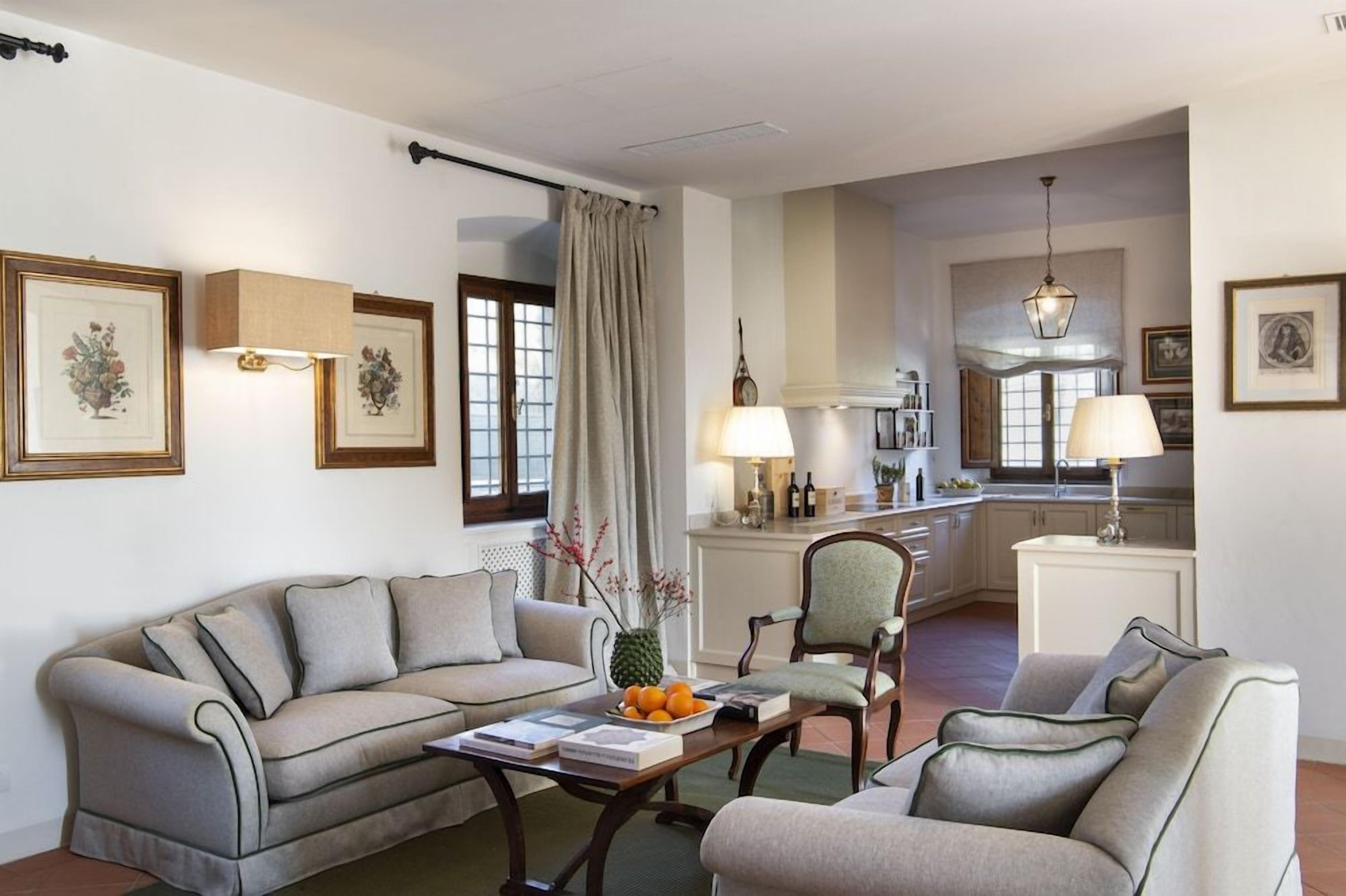 A living room with a sofa, armchairs, a coffee table with books and fruit, and a kitchen in the background.