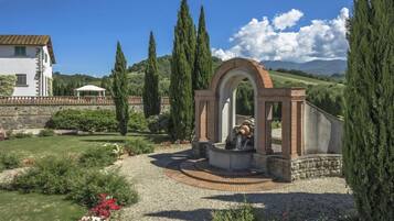 Un jardin avec une fontaine, un bâtiment blanc et de grands cyprès.