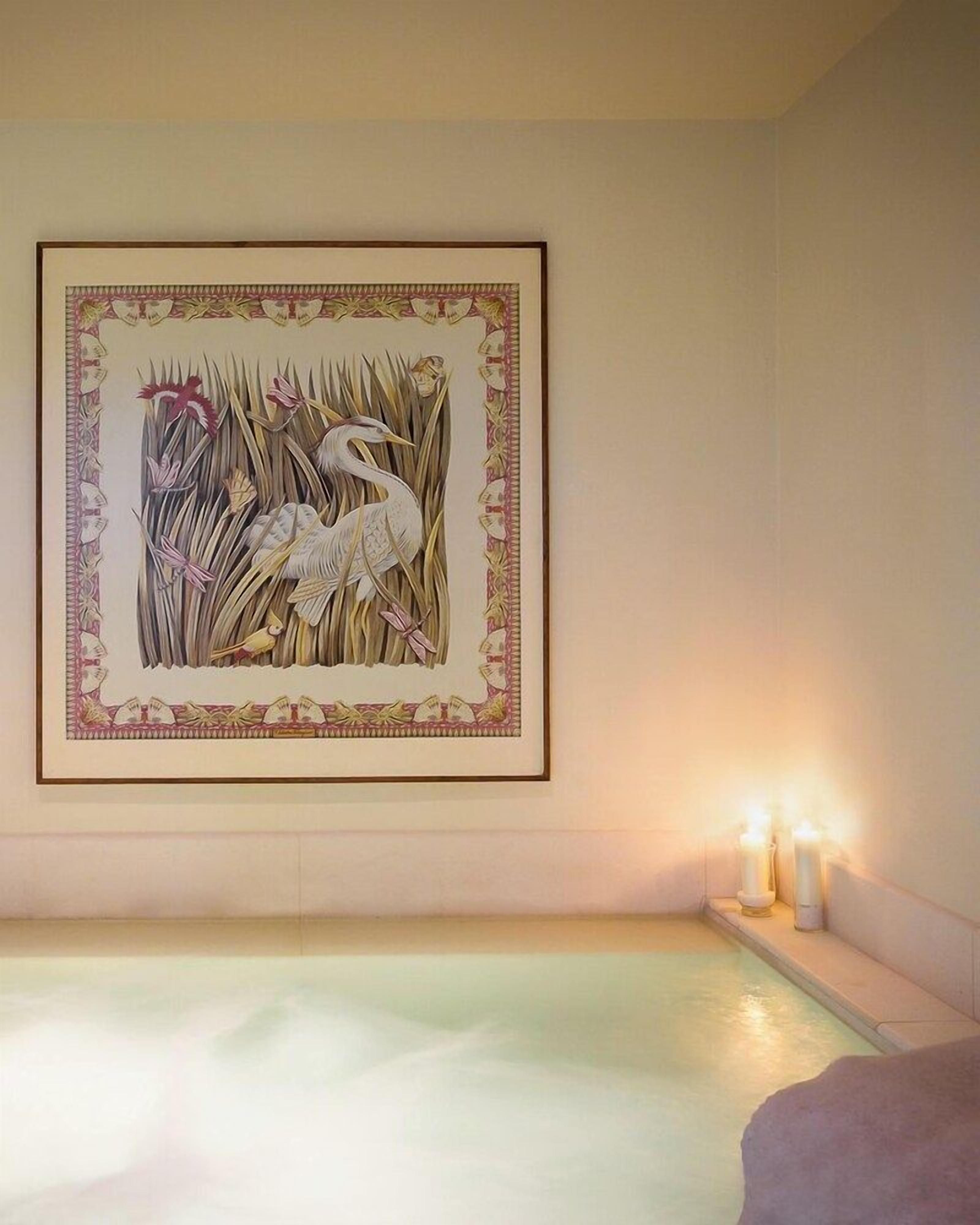 Un cuarto de spa con una obra enmarcada de pájaros y juncos en la pared, una bañera con velas sobre una mesita auxiliar y una fuente de agua visible.