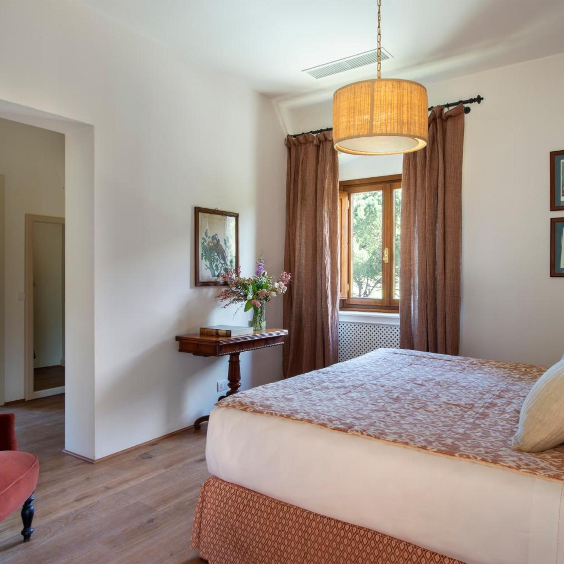Ein Schlafzimmer mit einem Bett, einem Nachttisch mit Blumen in einer Vase, einem Fenster mit Vorhängen und einer Pendelleuchte.