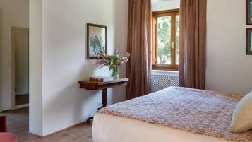 Ein Schlafzimmer mit einem Bett, einem Nachttisch mit Blumen in einer Vase, einem Fenster mit Vorhängen und einer Pendelleuchte.