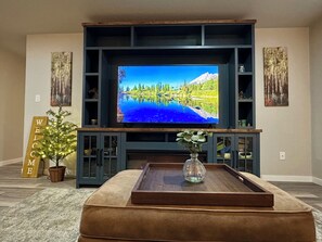 Ein Wohnzimmer mit einem großen Flachbildfernseher, der an einer dunklen Holz-Unterhaltungseinrichtung angebracht ist, einem bequemen braunen Hocker, einer kleinen Topfpflanze und einem Willkommensschild.