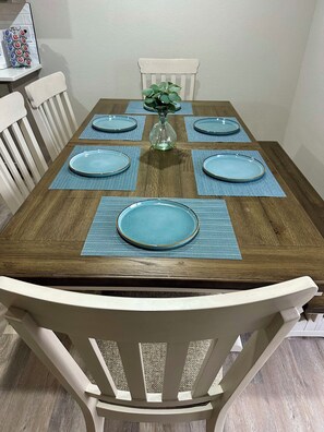 Mesa de jantar posta com pratos azuis e jogos americanos, um vaso com folhagem verde e uma cadeira branca.